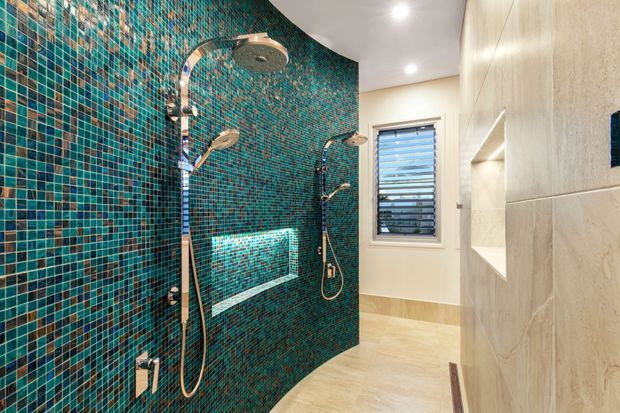 Contemporary walk-in bathroom shower featuring a curved wall clad in small iridescent teal and blue mosaic tiles, with dual chrome shower fixtures and a built-in tiled bench. Pale large-format wall tiles, a light tiled floor, recessed niches, and a louvred window create a clean, bright and refined finish.