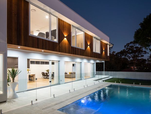 Twilight exterior view of a contemporary two-storey home with a projecting upper level clad in dark vertical timber, extensive glazing, warm wall lighting, and a ground-level covered terrace opening onto a swimming pool. The pool deck has frameless glass balustrading and pale paving, while the illuminated interior is visible through sliding glass doors.