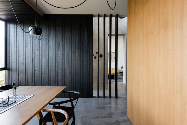 Contemporary dining area with a timber dining table and sculptural chairs, a dark vertically panelled feature wall, polished grey concrete flooring, and a timber-lined wall or built-in joinery. Slim black-framed glazed openings and a dramatic black linear pendant add an industrial-modern character, while a screened doorway leads toward another living space.
