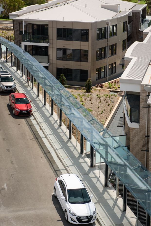 Elevated exterior view of a contemporary multi-storey building complex with curved dark-brick and light-panel facades, extensive glazing, flat roofs, balconies, landscaped areas, and a distinctive covered glass pedestrian walkway along the street. Vehicles are visible on the adjacent access road.