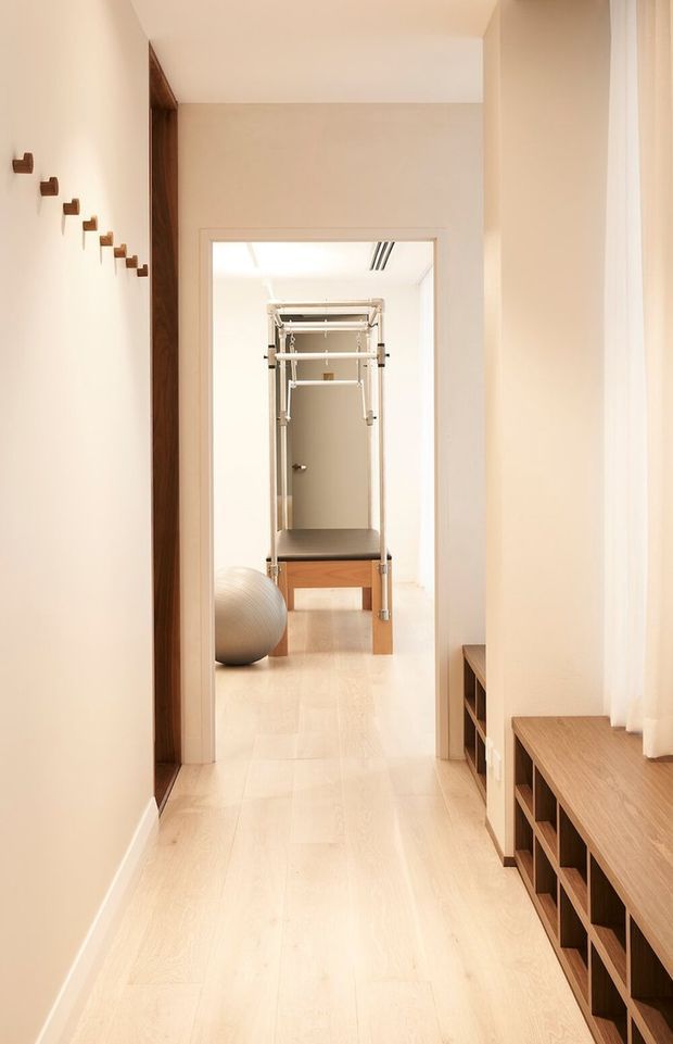 Bright, narrow residential hallway with pale timber flooring, warm white walls, built-in dark timber bench and open shelving along the right side, and sheer curtains over a large window. A doorway at the end opens to a compact home gym containing a wall-mounted exercise apparatus and exercise ball. Small dark timber wall hooks form a diagonal arrangement on the left wall.