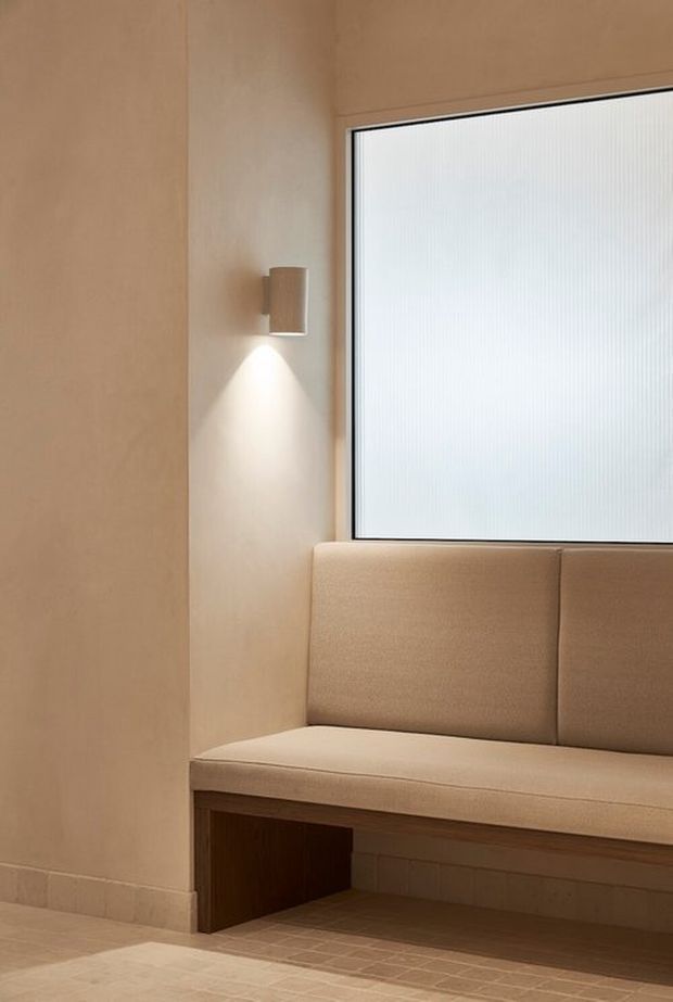 A minimalist built-in upholstered bench occupies a warm beige interior alcove beneath a large frosted or textured window. Light timber-toned wall panels, a dark timber bench base, pale tiled flooring, and a simple wall sconce create a calm, softly lit seating nook.