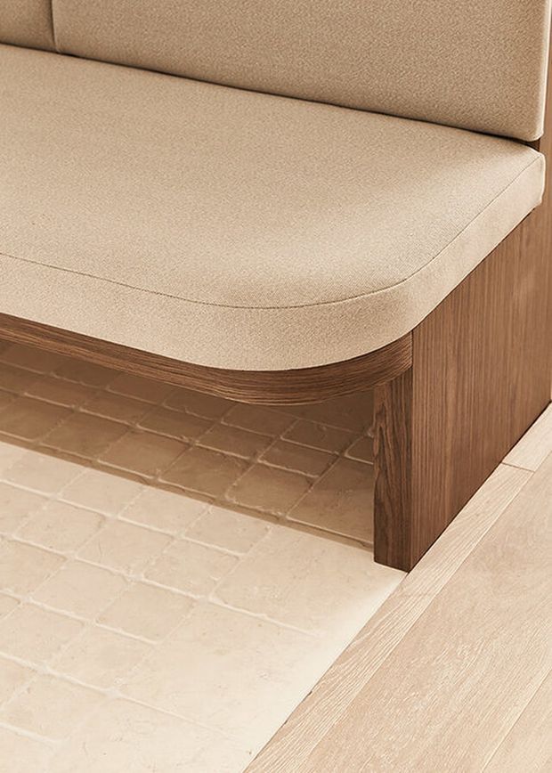 Close-up detail of a built-in upholstered bench or banquette with rounded cream fabric cushions, a warm medium-to-dark timber base and side panel, and pale textured tiled flooring beside light timber flooring. The image emphasizes the curved upholstery, joinery, and material transitions.