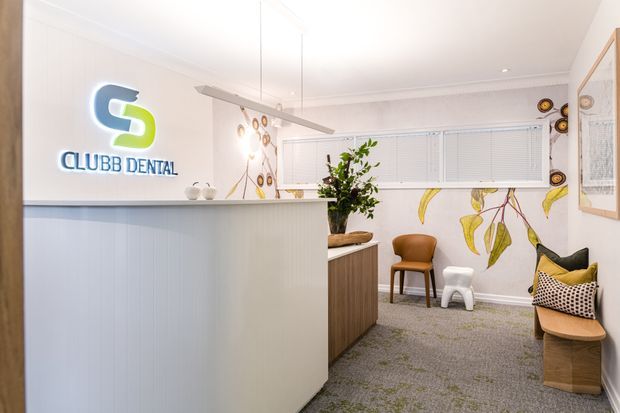 A bright contemporary dental clinic reception and waiting area. The foreground features a large white vertically panelled reception desk with an illuminated dental logo, while the waiting area contains timber seating, a small white stool, potted greenery, patterned cushions, carpet flooring, and botanical wall artwork. White venetian blinds and suspended linear lighting contribute to the clean commercial interior.