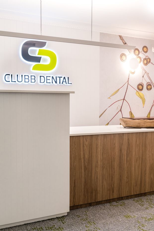 A bright contemporary dental clinic reception area featuring a white vertically panelled reception partition with illuminated branding, a wood-finished reception counter with a white benchtop, patterned carpet, and an illustrated botanical feature wall with decorative lighting and a timber bowl.