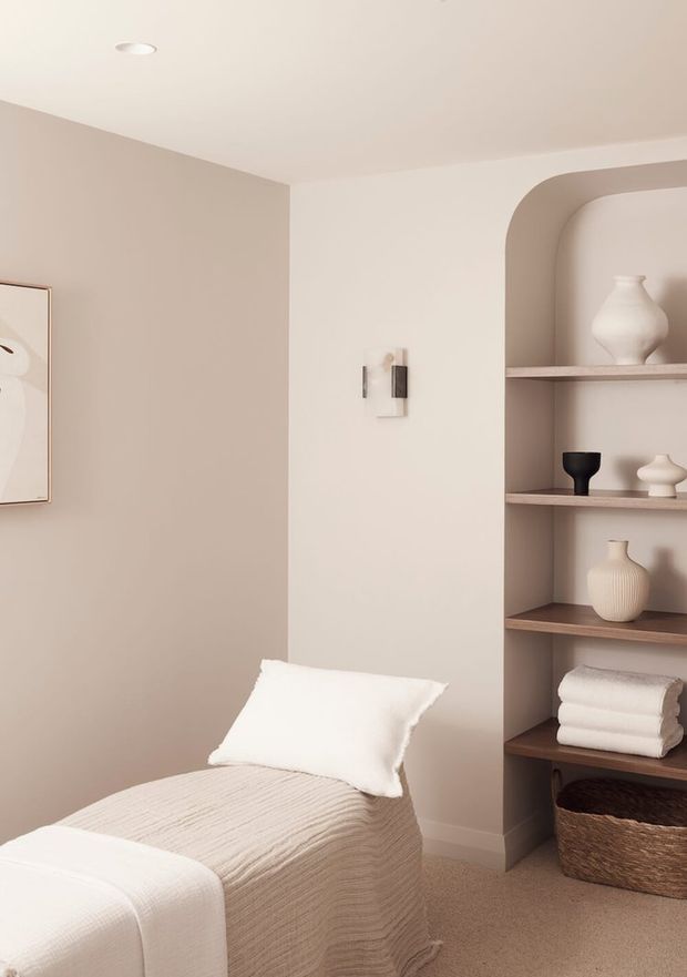 A softly styled bedroom corner featuring a single bed with white textured bedding and pillow, warm beige walls, and a built-in arched shelving niche. The shelves display neutral ceramic vessels, folded towels, and a woven basket, creating a calm minimalist interior.