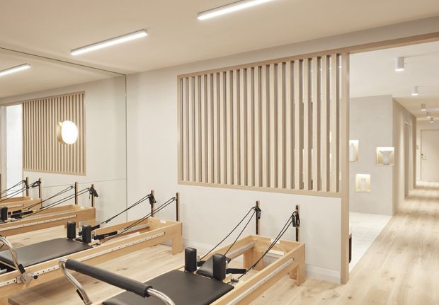 Bright, clean Pilates studio with multiple wooden reformer machines arranged in rows. The room features pale timber flooring, white walls and ceiling, linear recessed lighting, a large mirrored wall, and a decorative vertical timber slat screen separating the studio from a softly lit corridor and display niches.
