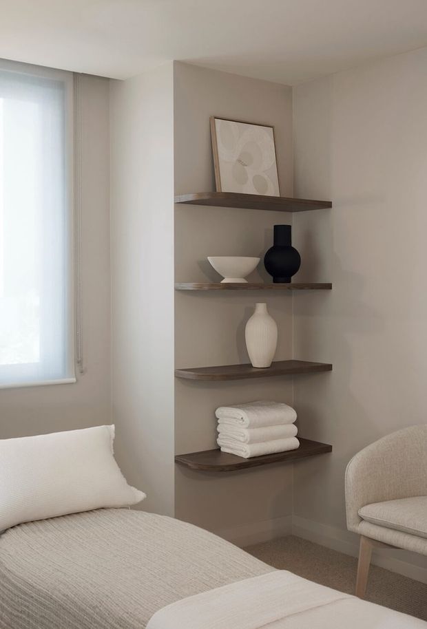 A calm, neutral-toned bedroom corner featuring a neatly made upholstered bed, a softly textured upholstered armchair, sheer window coverings, and a recessed wall niche with four dark timber floating shelves. The shelves display minimalist ceramics, artwork, and folded white towels. The room has soft beige walls, carpeted flooring, and gentle natural light.