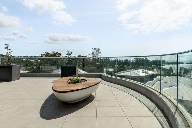 A spacious rooftop terrace with large pale stone or concrete-look paving, curved clear glass balustrades, and a sculptural oval outdoor table or planter in the centre. Large planters line the perimeter, with expansive suburban views beyond and a bright open sky.