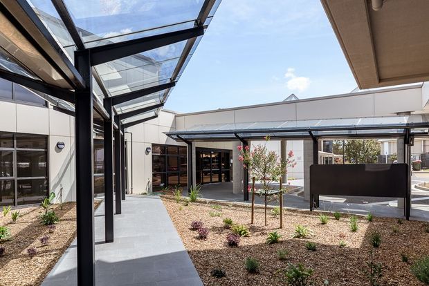 A contemporary single-storey courtyard viewed from within a building complex, featuring white panelled walls, dark metal-framed glazing, a broad paved walkway, extensive overhead glass canopies, and low-maintenance landscaped garden beds with young trees and shrubs. The open courtyard is bright and airy, with a covered outdoor seating area visible at the far side.