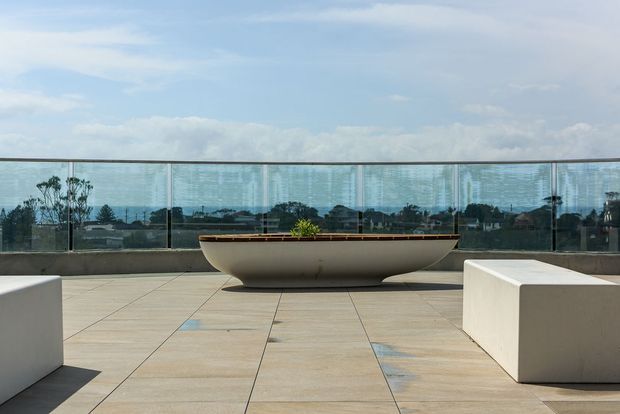 A contemporary rooftop terrace or balcony with large beige tiled paving, a frameless glass balustrade, and minimalist white built-in bench seating. A long sculptural planter sits centrally, with a distant urban and coastal-looking view beyond the railing under a bright sky.