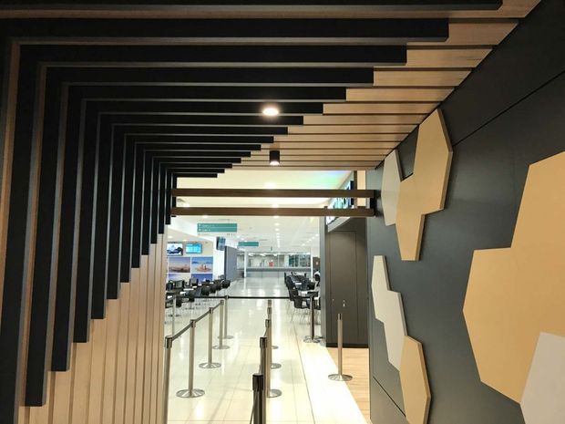 A contemporary commercial interior, likely an airport or transport terminal, viewed through a dramatic corridor-like passage. Repetitive black overhead beams create a strong linear rhythm over light timber slats, while dark wall panels feature large geometric beige acoustic or decorative forms. The passage opens into a bright polished public waiting and queuing area with stanchions, seating, signage, and service counters.
