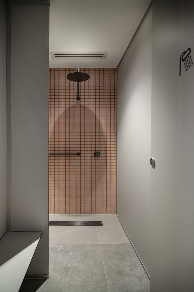 A compact contemporary walk-in shower with a warm blush-pink small-format tiled feature wall, dark metal overhead shower and controls, pale grey floor tiles, and smooth light-grey walls. A linear floor drain and recessed ceiling ventilation add to the clean, minimalist fit-out.