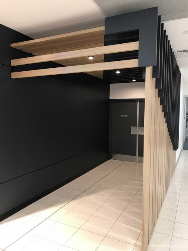 Modern commercial interior corridor or entry zone featuring a bold black feature wall, light timber slatted screening, projecting timber ceiling fins, dark acoustic ceiling panels, recessed lighting, and pale tiled flooring. A dark lift door is visible at the end of the passage.