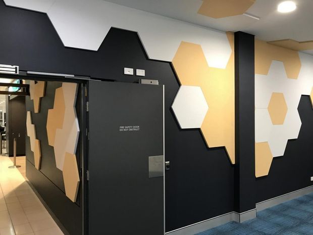 A commercial interior corridor or entry area with a bold geometric feature wall and dark door. The walls are primarily matte black with irregular angular edges and layered hexagonal and polygonal panels in white, beige, and tan. A blue carpet, tiled flooring, metal skirting, exposed services, and a contrasting illuminated corridor are visible. The space has a graphic, contemporary workplace or public-building fit-out.