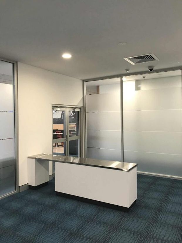 A clean, modern commercial reception area with a white rectangular reception desk featuring a dark stone or solid-surface countertop and black plinth. The space has frosted glass partition walls and doors with horizontal clear bands, white walls and ceiling, recessed lighting, exposed HVAC grilles, security fixtures, and dark blue patterned carpet. The room appears minimal and functional, likely an office lobby or workplace entry.