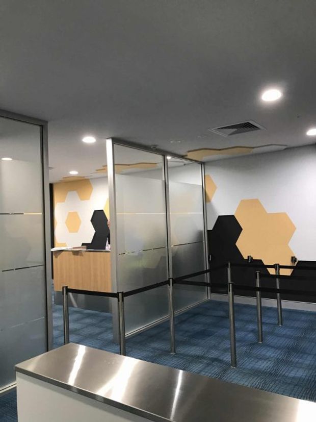 A contemporary commercial interior, likely a reception or service waiting area, with frosted glass partitions in dark metal frames, queue barriers, blue carpet, a timber reception counter, stainless-steel foreground counter, recessed ceiling lights, and graphic black and ochre geometric wall panels.