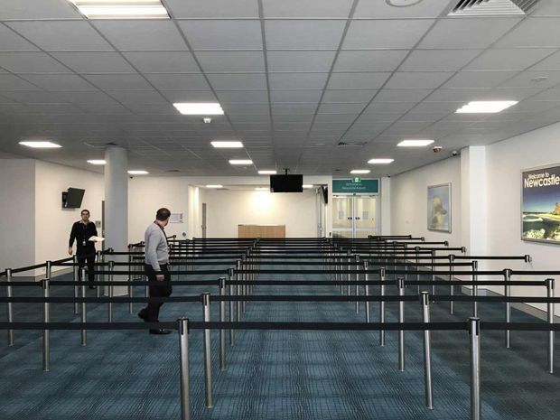 A large, utilitarian airport or transport-terminal queuing area with extensive black metal crowd-control barriers arranged in lanes across a blue carpeted floor. The space has white walls, round structural columns, a suspended acoustic tile ceiling with recessed fluorescent lighting and ventilation grilles, wall-mounted signage and artwork, and a service or security checkpoint at the far end. A few people are present, but the primary subject is the commercial circulation and waiting space.