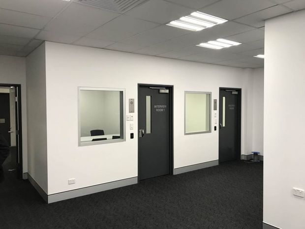 A clean institutional or commercial interior corridor with white partition walls, dark carpet flooring, black doors labeled as interview rooms, internal observation windows, and a suspended acoustic ceiling with fluorescent lighting. The space appears functional and professionally fitted out, with a neutral monochrome palette.