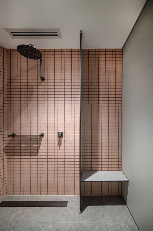 A contemporary walk-in shower with a full-height pink square-tile feature wall, dark grout, matte black rainfall shower and controls, a slim glass shelf, linear floor drain, and a built-in white bench. Large grey floor tiles and neutral walls create a clean, modern bathroom interior.