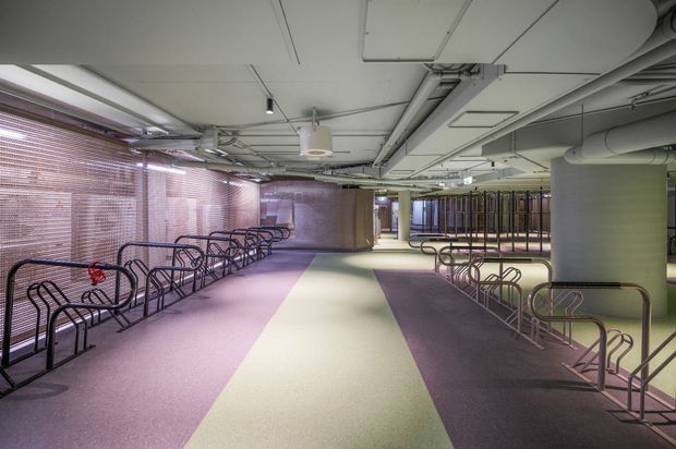 A large, brightly lit interior bicycle parking facility with rows of metal bike racks, a broad central circulation path, exposed services and ductwork, structural columns, and translucent screened glazing along the perimeter. The floor uses broad bands of muted purple, pale green, and dark grey.