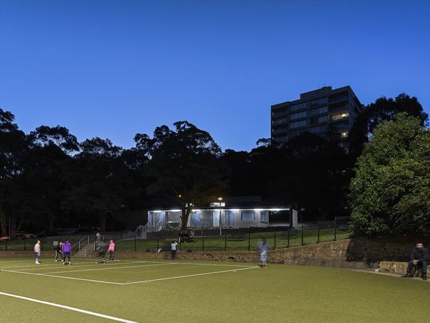 A dusk exterior view of a fenced outdoor sports court, likely a tennis court, with players on the artificial turf. A low, brightly lit clubhouse or sports facility sits behind the court, surrounded by mature trees, with a multi-storey apartment or commercial building visible on the elevated site in the background.