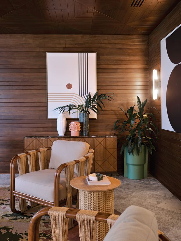 Warm, richly layered lounge or sitting area with dark timber wall and ceiling panelling, a textured stone or concrete-look floor, woven cane and rattan armchairs, a round woven side table, sculptural artwork, and abundant indoor plants. The space has a refined retro-tropical feel with integrated wall lighting and timber storage cabinetry.