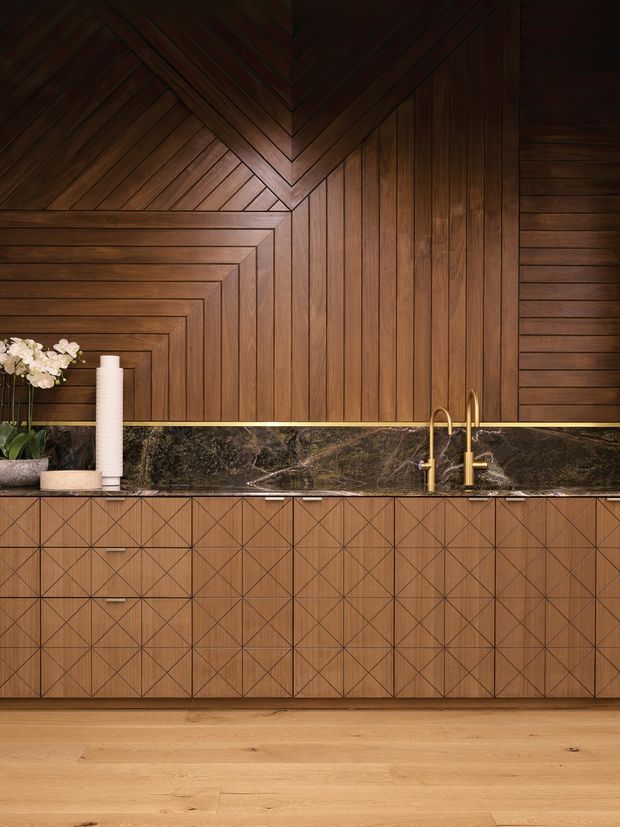A richly detailed timber kitchen or bar joinery installation featuring geometric patterned wood cabinetry, vertical and chevron wall panelling, a dark veined stone benchtop and splashback, and brushed brass tapware. Warm timber flooring and subdued lighting create a refined, moody atmosphere.
