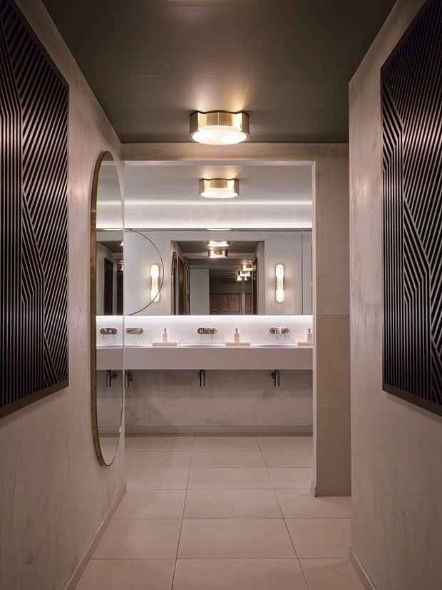 A refined, dimly lit bathroom vanity area viewed down a narrow tiled passage. The space features multiple wall-mounted basins with chrome tapware, a large mirrored wall, warm integrated and pendant-like lighting, pale tiled flooring and walls, curved mirror detailing, and dark geometric slatted feature panels on both side walls.