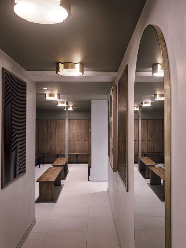 A refined contemporary changing-room or locker area viewed from a corridor, featuring warm wood locker cabinetry, long timber benches, pale tiled flooring, dark textured wall panels, a large arched mirror, and circular flush-mounted ceiling lights. The space is narrow but extends into a larger locker zone through the opening.