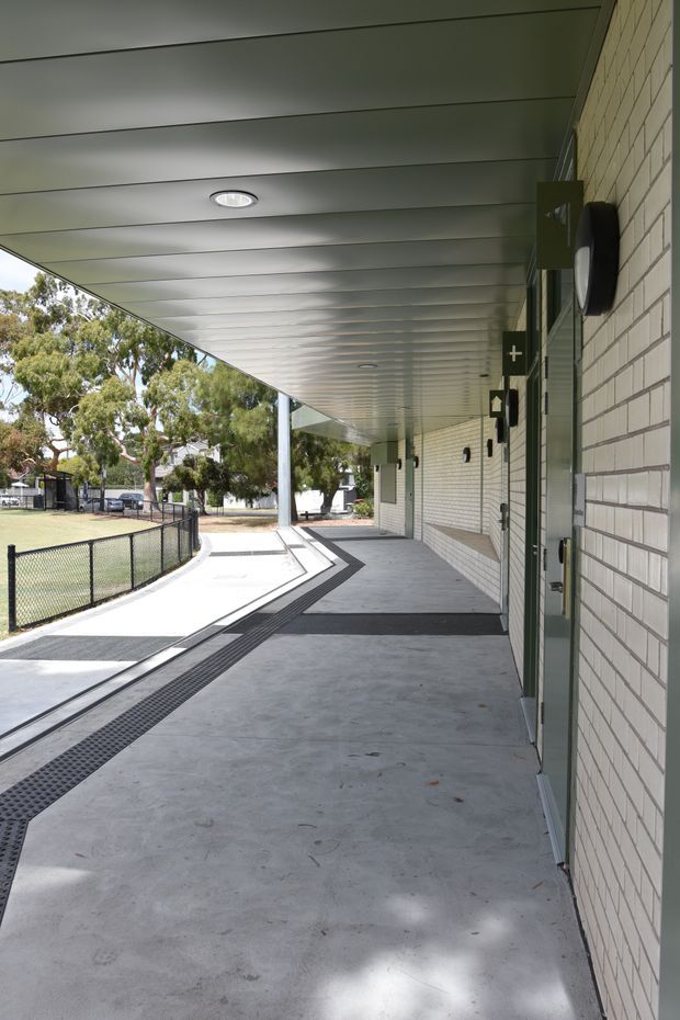 A covered exterior walkway beside a light brick building, with a broad smooth concrete floor, continuous grey metal soffit ceiling with recessed lights, wall-mounted fixtures and doors, and a black grated drainage channel following the path. A chain-link fence and landscaped grounds are visible to the side. The setting has a practical institutional or school-like character.