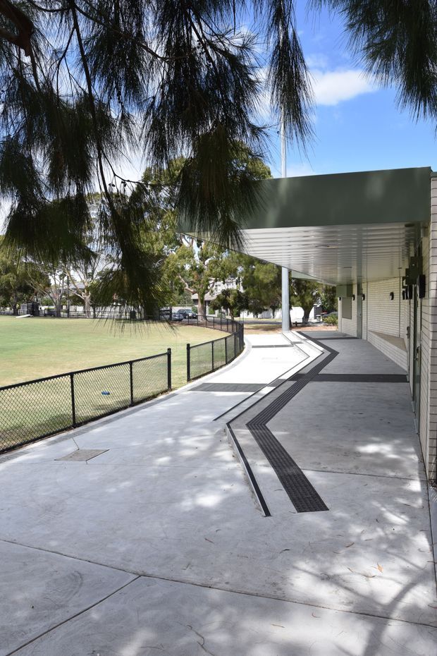 Exterior view of a contemporary low-rise building beside a fenced grass recreation area. A broad concrete walkway curves along the building beneath a projecting canopy, with dark tactile paving strips and a light brick wall. Mature trees provide shade, and the scene has a clear, institutional or community-facility character.