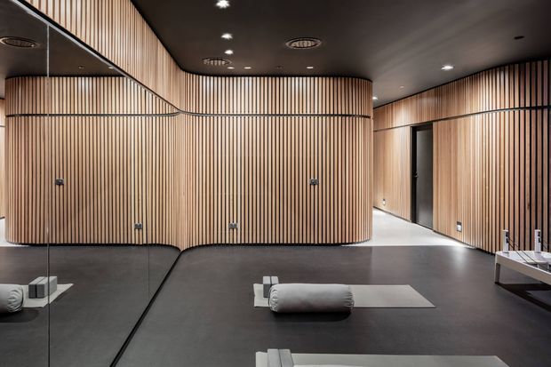 A refined contemporary wellness or yoga studio with a dark matte floor, soft neutral yoga mats and bolsters, and extensive curved walls clad in vertical timber battens. Full-height mirrors line the left wall, while recessed downlights and circular ventilation grilles punctuate the dark ceiling. The slatted timber treatment creates a warm, rhythmic interior around the open exercise area.