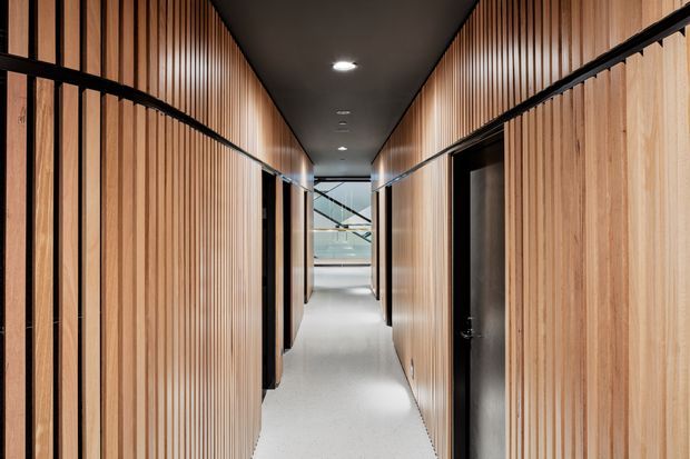A narrow modern interior corridor lined on both sides with vertical timber slat wall panelling and black-framed door openings. The passage has a smooth dark ceiling with recessed downlights, a light grey floor, and glazing at the far end revealing a bright open area beyond. The composition emphasises the rhythm of the timber battens and clean, contemporary detailing.