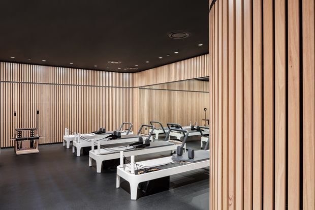 A modern Pilates studio or fitness room with several white reformer machines arranged in rows. The space features full-height vertical timber slat wall panelling, a large wall mirror, dark flooring, and a matte dark ceiling with recessed lighting. The restrained material palette and integrated lighting create a refined, calm, contemporary fitness environment.