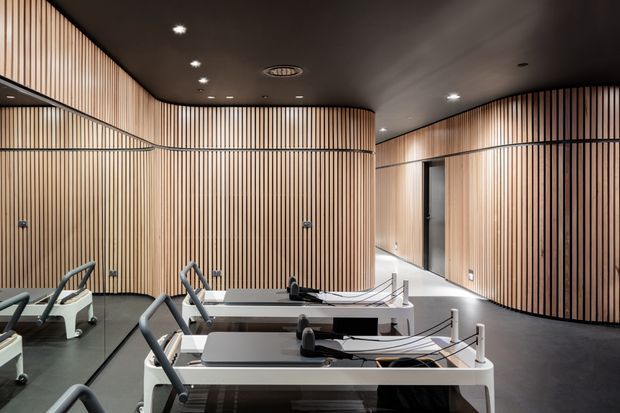A refined commercial fitness studio with several modern reformer-style exercise machines arranged in rows. The space features extensive curved walls clad in vertical timber slats, a dark smooth ceiling with recessed spotlights, dark flooring, and a large mirror along the left wall. A corridor and doorway are visible beyond the curved partition.