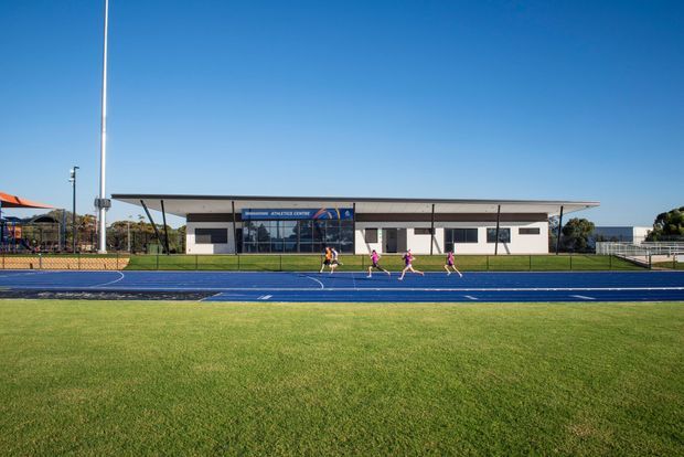 A wide exterior view of a modern single-storey athletics centre beside a blue running track and expansive grass sports field. The building has a long, low rectangular form, a shallow projecting roof, dark-framed glazing at the central entrance, and a mix of light rendered walls and dark horizontal window bands. Fencing, sports lighting, and athletes reinforce the institutional sporting setting.