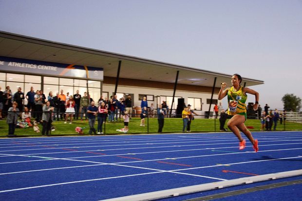Exterior photograph of an athlete sprinting on a blue synthetic running track beside a modern athletics centre. Spectators stand along the track, with the low, broad building and glazed entrance visible behind them.