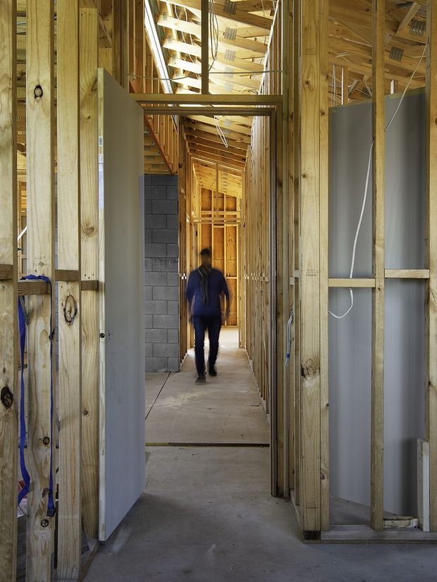 A construction-stage interior viewed down a long corridor, with exposed timber wall and roof framing, unfinished grey wall linings, visible electrical wiring, concrete blockwork, and a bare concrete floor. A person stands in the distance, emphasizing the scale and depth of the space. The image documents the building structure before completion rather than a finished room.