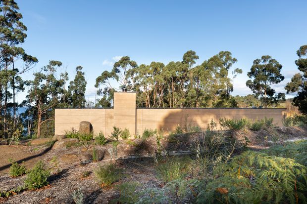 Low, minimalist masonry building set within a native landscaped setting. The warm tan rendered or masonry walls form a long horizontal volume with a raised rectangular chimney or tower element and a small arched opening. Eucalypt trees and drought-tolerant planting surround the structure, under clear daylight.