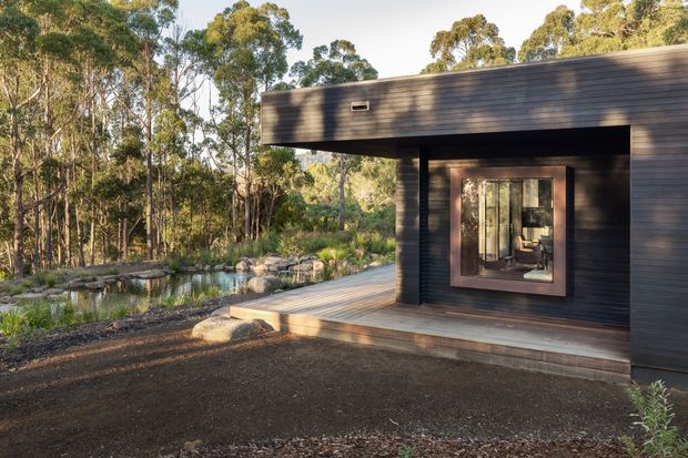 Exterior view of a compact modern pavilion or cabin set within a wooded landscape. The building has dark horizontal timber cladding, a flat projecting roof, a large glazed opening with a timber-framed window, and a timber deck extending toward a natural pond edged with rocks and planting. The setting is quiet and integrated with native woodland.