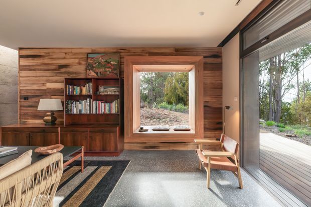 Warm, mid-century-inspired living room with a timber-lined feature wall, integrated dark-wood bookcase and cabinetry, and a deep-set window overlooking trees. A large glazed sliding door opens onto a timber deck, bringing abundant natural light into the compact sitting area. The room includes a woven sofa, low timber coffee table, lounge chair, floor lamp, bookshelves, and a terrazzo-like dark floor with a geometric rug.