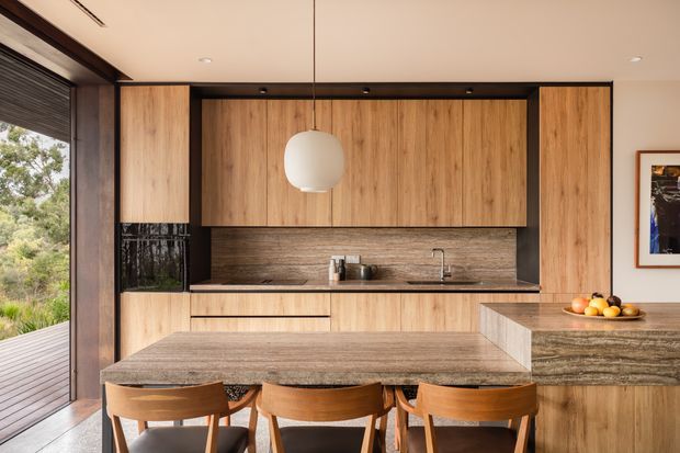 A warm, contemporary open-plan kitchen and dining area with extensive timber cabinetry, a stone island bench, matching stone benchtops, and a textured stone backsplash. Three timber dining chairs sit at the island beneath a spherical pendant light. Full-height glazing and a timber deck connect the kitchen to a leafy outdoor setting.