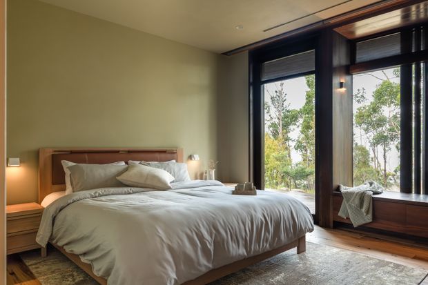 A warm, refined bedroom with a low-profile timber bed, layered white bedding, bedside tables, and soft wall lighting. Olive-green walls contrast with dark timber-framed floor-to-ceiling windows and integrated window seating, overlooking trees. The room has timber flooring, a neutral rug, and a calm natural atmosphere.