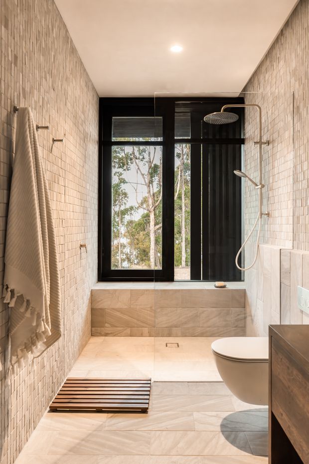 A narrow contemporary bathroom with a walk-in shower, freestanding-style wall-mounted shower fixtures, toilet, and a deep tiled window ledge. Small pale rectangular wall tiles cover both side walls, while larger light stone-look tiles with diagonal veining cover the floor and lower ledge. Black-framed windows provide views to trees, and a wooden slatted bath mat and neutral towel add warmth.