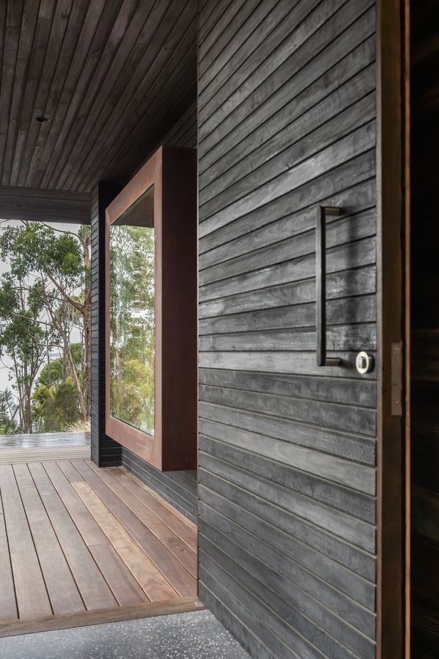 A close architectural view of a dark timber-clad entry porch, with horizontal weathered wood boards covering the wall and ceiling, a timber deck, a large framed window, and a simple vertical metal door handle. The covered threshold opens toward a leafy wooded setting, creating a restrained, natural cabin-like entrance.