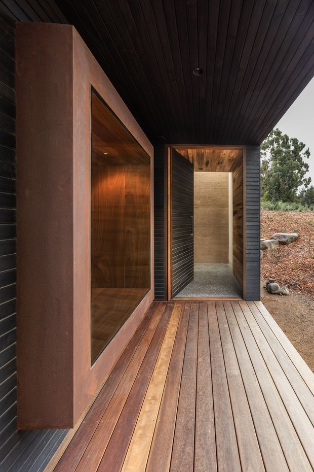 A dark, minimalist architectural entry or outdoor passage with a covered timber deck. The facade combines black horizontal cladding, weathered rust-toned metal, warm timber-lined recesses, and a concrete-finished open doorway. A dark timber soffit extends overhead, while sparse landscaped ground and rocks are visible beyond.