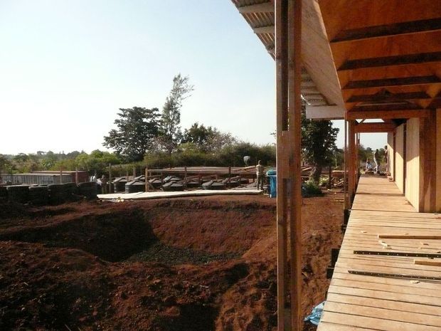 Exterior view of a timber-clad building or structure under construction, featuring a raised timber walkway or deck with a deep covered verandah, exposed timber posts and beams, and corrugated roofing. The surrounding reddish earth, construction materials, and partially completed landscape indicate an active building site.