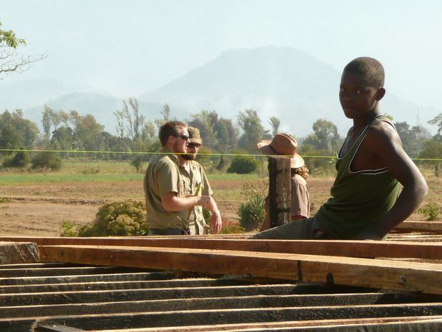An outdoor construction scene with workers standing beside timber framing or formwork. A rural landscape with trees and a hazy mountain is visible in the background, while stacked wooden beams and dark structural members dominate the foreground. The image has a documentary, warm daylight character.