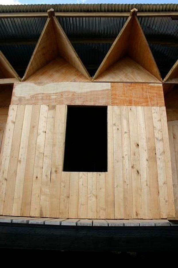 A close-up of a small timber-clad structure under a corrugated metal roof. The facade is made from vertical unfinished timber boards, with a dark rectangular doorway or window opening and a distinctive row of triangular plywood roof or gable elements beneath the roofline. The image appears to document an in-progress or utilitarian construction.